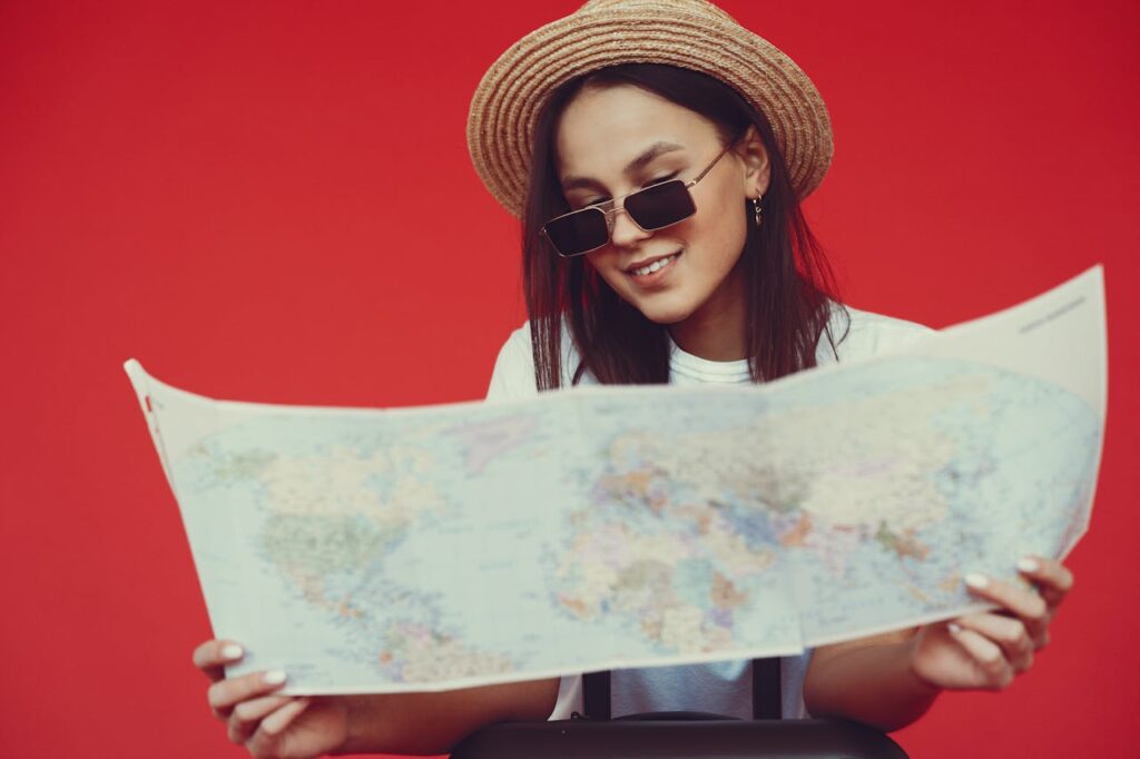 Smiling young woman in stylish hat and sunglasses choosing destination on paper map while standing with luggage on red background